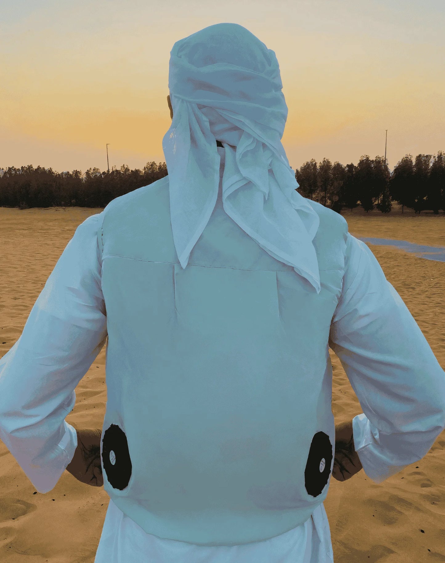 Person wearing a white outfit and Naseem cooling vest equipped with two black fans attached from the backside in a desert setting during sunset.